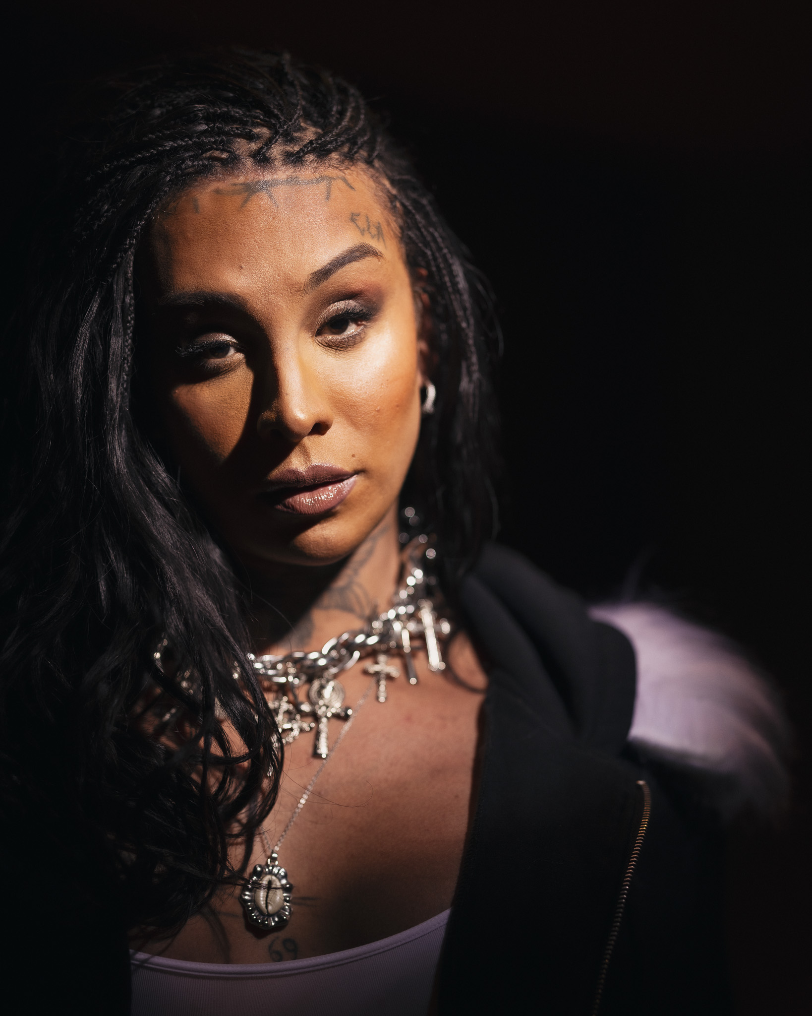 A portrait of Linn da Quebrada. The background is black. A light shines on Linn da Quebrada from the top right of the photo.